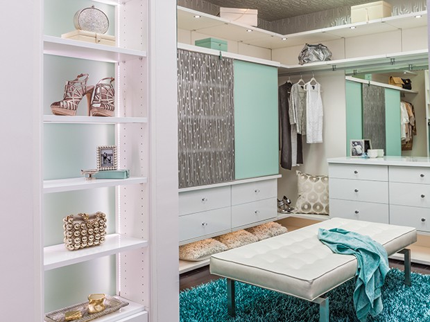 A walk in closet in a light wood grain finish with blue and green accents, open shelving, cabinets and built in dresser by California Closets